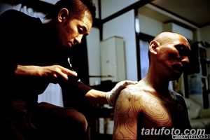Фото процесса нанесения татуировки в стиле трайбл на плечо. Мастер тату работает над рисунком, клиент сидит в кресле.