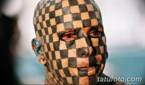 Крупный план лица мужчины, покрытого черно-белой шахматной татуировкой. Его губы слегка приоткрыты, а взгляд направлен прямо в камеру.