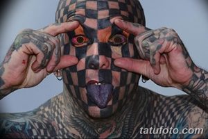 Фото тату: мужчина с татуировкой в виде шахматной доски на лице, высунутым языком и руками, обрамляющими глаза. Яркие цвета и провокационное выражение лица.