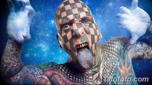 Фотография мужчины с полным татуированным телом, включая лицо, покрытое черно-белой клеткой. Выражение лица агрессивное, язык высунут.