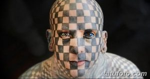 Фото тату: лицо мужчины полностью покрыто черно-белой шахматной татуировкой. Мужчина смотрит в камеру.