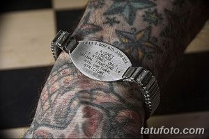 Фото тату: рука мужчины, покрытая татуировками, с металлическим браслетом, содержащим медицинскую информацию.
