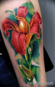 Фото реалистичной татуировки с изображением цветка ириса на руке. Яркие красные и оранжевые оттенки, детализированные лепестки и листья.