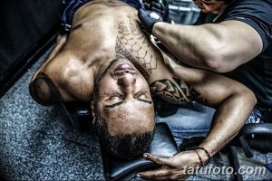Фотография мужчины с татуировкой в стиле трайбл на груди и плечах. Виден процесс нанесения татуировки.