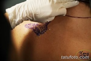 Фотография процесса переноса эскиза татуировки бабочки на лопатку девушки. Мастер использует трафарет и маркер.