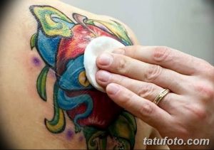 Фото процесса обработки свежей татуировки: мастер протирает яркую татуировку цветка на плече клиента ватным диском.