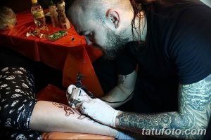 Фото процесса нанесения татуировки на ногу клиента. Мастер тату с бородой и длинными волосами сосредоточенно работает над рисунком.