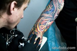 Фото процесса нанесения яркой и красочной татуировки на руку клиента. Мастер тату в черной одежде сосредоточенно работает над рисунком.
