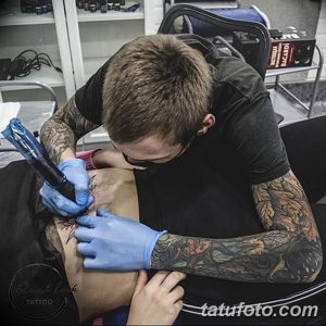Фото процесса нанесения цветной татуировки на спину клиента. Мастер тату в черной футболке сосредоточенно работает над рисунком.
