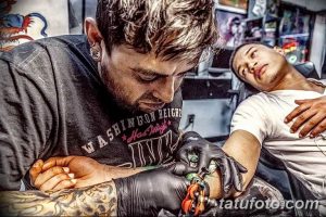 Фото процесса нанесения татуировки на руку клиента. Мастер тату в черной футболке сосредоточенно работает над рисунком, используя тату-машинку.