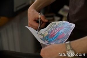 Фото тату процесса: мастер вырезает эскиз татуировки ножницами. Видна рука мастера, эскиз и рука клиента.
