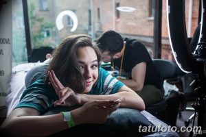 Фото тату процесса: девушка сидит в кресле во время сеанса татуировки, улыбается и показывает знак победы. Мастер работает над татуировкой за ее спиной.