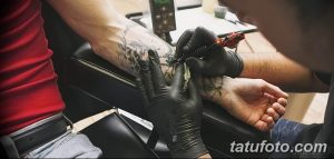 Фото тату процесса: мастер в черных перчатках наносит татуировку на руку клиента. Видна тату-машинка и частично уже выполненный рисунок.