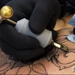 Фото тату процесса нанесения татуировки розы на спину: рука мастера в черных перчатках, тату-машинка, контурный рисунок розы. Детализированный снимок.