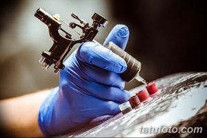 Фото тату процесса нанесения татуировки: рука мастера в синих перчатках, тату-машинка, баночки с краской. Детализированный снимок работы.