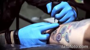 Фото процесса нанесения яркой татуировки с использованием цветных чернил на предплечье. Мастер в голубых перчатках работает над рисунком.