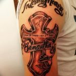 Фото тату креста с именем и датами на плече. Черно-белый рисунок в стиле олдскул, посвященный памяти ушедшего человека.