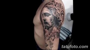 Фото тату на плече с изображением лика Иисуса Христа. Реалистичный черно-серый рисунок с декоративными элементами.