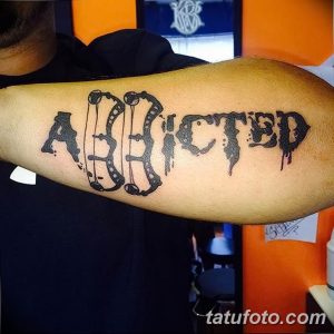 Фото тату: Цветная татуировка надписи 'Addicted', выполненная в стиле леттеринг с эффектом брызг краски, расположена на предплечье.