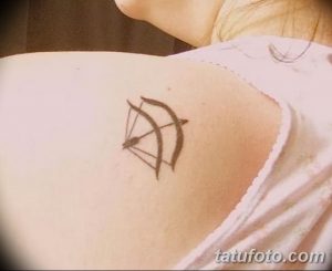 Фото тату: Маленькая черно-белая татуировка лука и стрелы, расположенная на плече. Изящный эскиз тату с тонкими линиями.