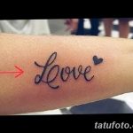 Фото тату надписи 'Love' на женском запястье, выполненной черными чернилами. Небольшое изображение сердца дополняет композицию.