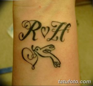 Фотография татуировки на запястье, изображающей буквы 'R' и 'H', соединенные сердцем, и двух голубей. Черно-белый рисунок татуировки.