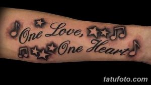 Фото тату на предплечье с надписью 'One Love, One Heart' и музыкальными нотами, украшенными звездами и брызгами.