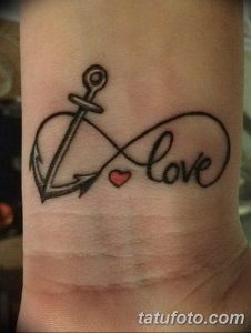 Фото тату надписи 'Love', переплетенной с изображением якоря и символом бесконечности, на запястье. Черный цвет, изящный стиль.