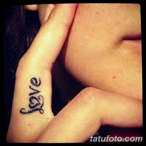 Фотография небольшой татуировки с надписью 'love' на боковой стороне пальца. Черный контур, изящный шрифт, женственный дизайн.