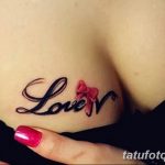 Фото тату надписи "Love" с красным бантиком, расположенной на груди. Элегантный шрифт и нежный дизайн делают татуировку женственной и романтичной.