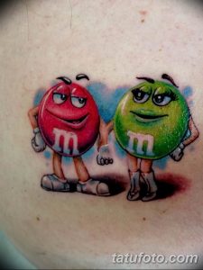 Фото тату двух M&M's – красного и зеленого – с милыми глазками и улыбками, выполненное в ярком, мультяшном стиле. Расположено на плече.
