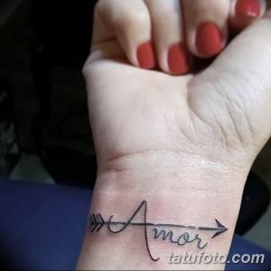Фото татуировки на запястье. Изображена надпись 'Amor' со стрелой, пронзающей слово. Черно-белый рисунок татуировки, выполненный в минималистичном стиле.