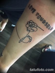Черно-белое фото татуировки на предплечье с надписью 'Love Yourself' и стилизованным изображением сердца, выполненным тонкими линиями.