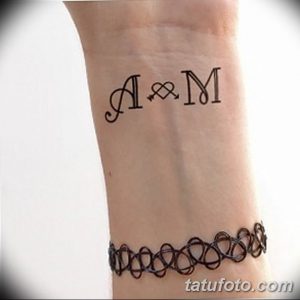 Фото татуировки с инициалами 'A & M', соединенными символом амперсанд. Черно-серый рисунок тату на запястье.