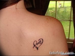 Фото тату на плече: надпись 'Love' стилизованная под форму сердца, выполненная черными чернилами.