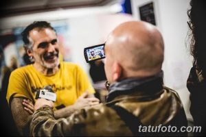 Фотография известного мастера тату, дающего интервью на тату-конвенции. Виден его довольный вид и интерес к нему со стороны журналиста.