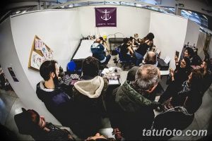 Фото тату-конвенции с множеством людей, мастеров и эскизов татуировок. Видна работа мастера над тату, интерес к рисункам и оживленная атмосфера.