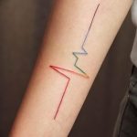Фото тату на предплечье, изображающее кардиограмму, линии которой окрашены в цвета радуги. Оригинальный и символичный рисунок.