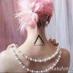 Фото тату: простой черный треугольник на затылке девушки с розовыми волосами. Минималистичный дизайн, геометрическая форма, стильный аксессуар.