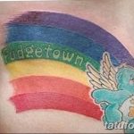 Фото татуировки на боку с изображением радуги и мультяшного медведя Care Bear с крыльями, выполненной в ярких цветах.
