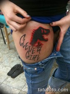 Фото тату на бедре: красный динозавр с надписью 'Clever Girl', выполненное в простом, мультяшном стиле.