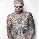 Фотография мужчины, полностью покрытого сложными черными татуировками, включая лицо, шею и тело. Изображение создает впечатление мрачного и загадочного образа.