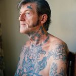 Фотография пожилого мужчины с татуировками в стиле олдскул на груди, руках и шее.