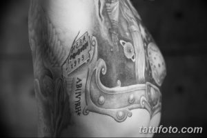 Фото тату на плече: детализированный черно-белый рисунок рыцарского шлема и свитка с надписью. Реалистичная татуировка с историческим мотивом.