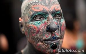 Фото татуировки на лице: мужчина с полностью татуированным лицом в ярких цветах, с пирсингом и необычным выражением лица.