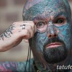 Фото тату: мужчина с полностью зататуированным лицом, рассматривающий что-то через лупу. Детализированный рисунок татуировки покрывает всю кожу лица.