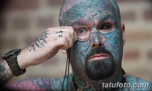 Фото тату: мужчина с полностью зататуированным лицом, рассматривающий что-то через лупу. Детализированный рисунок татуировки покрывает всю кожу лица.