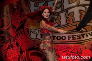 Фото модели с татуировками на тату-фестивале, одетой в красный наряд и с ярким макияжем. На заднем плане логотип фестиваля.