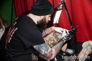 Фото тату мастера, работающего над татуировкой на теле клиента. Мужчина с бородой, сосредоточенно наносящий рисунок.