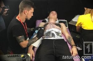 Фото тату: мастер делает татуировку мужчине, лежащему на кушетке. Видны инструменты для татуировки и процесс нанесения рисунка.